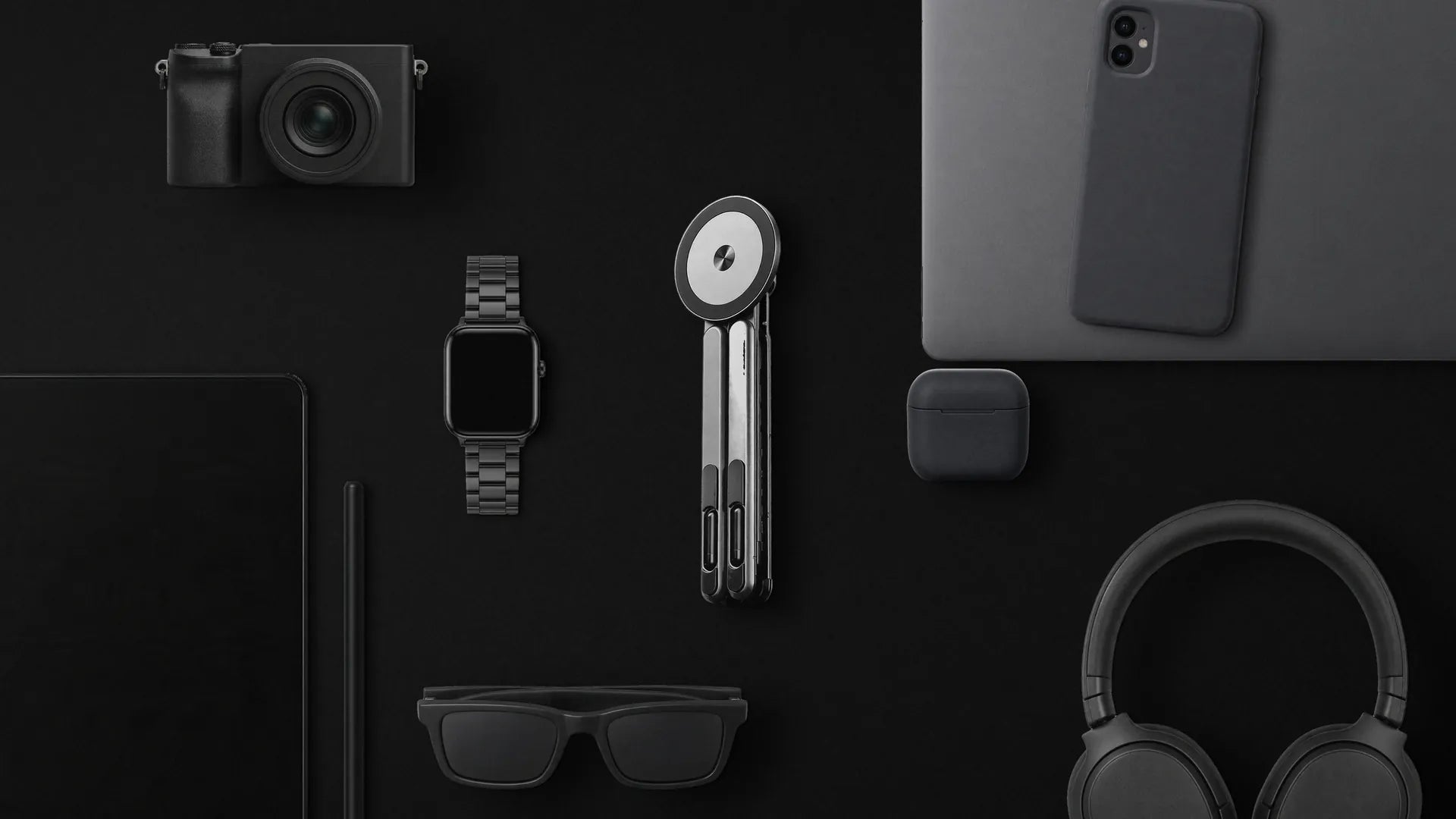 Vante Polaris gris plegado sobre una superficie negra mate, visto desde arriba, entre una cámara, un reloj, una tablet con un móvil, unos auriculares, unos cascos, unas gafas y una libreta